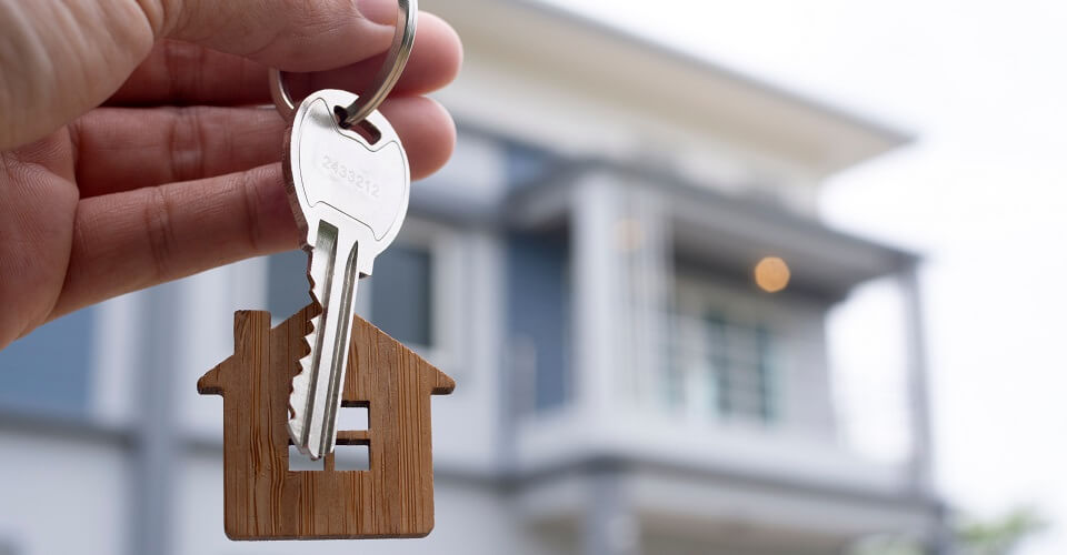 Hand holds a keychain with a key and model of a house and behind it a house