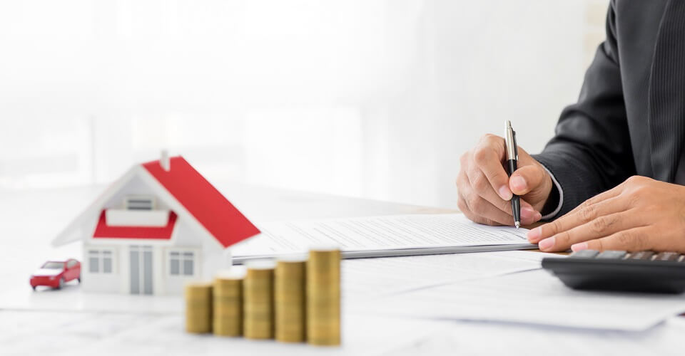 A model of a house next to coins and someone writing in a document with a pen